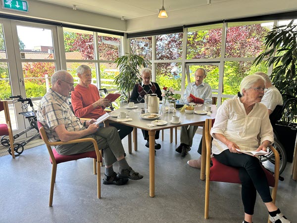 En gruppe ældre synger fællessange og bladrer i sangbøger ved et bord omgivet af grønne planter og store vinduer